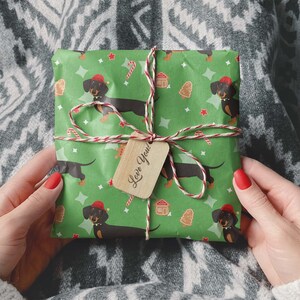 K&ouml;nnte beinhalten: Ein Geschenk, verpackt in gr&uuml;nem Papier mit einem Muster aus Dackeln mit Weihnachtsm&uuml;tzen, Zuckerstangen, Lebkuchenh&auml;usern und Weihnachtsb&auml;umen. Es ist mit Bindfaden und einem Holzanh&auml;nger mit der Aufschrift "Love You!" gebunden.
