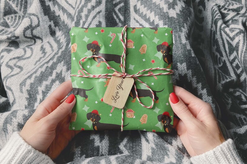 K&ouml;nnte beinhalten: Ein Geschenk, verpackt in gr&uuml;nem Papier mit einem Muster aus Dackeln mit Weihnachtsm&uuml;tzen, Zuckerstangen, Lebkuchenh&auml;usern und Weihnachtsb&auml;umen. Es ist mit Bindfaden und einem Holzanh&auml;nger mit der Aufschrift "Love You!" gebunden.