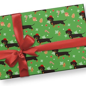 K&ouml;nnte beinhalten: Ein verpacktes Geschenk mit gr&uuml;nem Hintergrund und einem sich wiederholenden Muster aus schwarzen Dackeln mit roten Weihnachtsm&uuml;tzen, Lebkuchenm&auml;nnern, Zuckerstangen und Sternen. Ein rotes Satinband ist um das Geschenk gebunden.