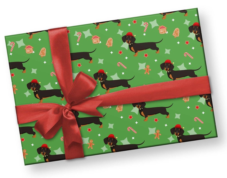 K&ouml;nnte beinhalten: Ein verpacktes Geschenk mit gr&uuml;nem Hintergrund und einem sich wiederholenden Muster aus schwarzen Dackeln mit roten Weihnachtsm&uuml;tzen, Lebkuchenm&auml;nnern, Zuckerstangen und Sternen. Ein rotes Satinband ist um das Geschenk gebunden.