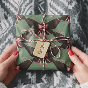 May include: A wrapped gift with a green background and a repeating black butterfly pattern. The gift is tied with a red and white string and a small wooden tag that says "Love You!".