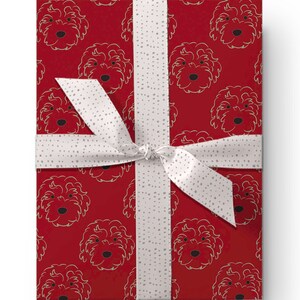 May include: A wrapped gift box featuring a red background with a repeating pattern of dog faces in a gold outline. The box is tied with a white ribbon with small gray polka dots. The ribbon is tied in a bow.