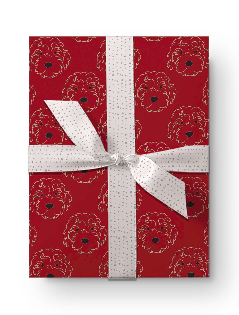 May include: A wrapped gift box featuring a red background with a repeating pattern of dog faces in a gold outline. The box is tied with a white ribbon with small gray polka dots. The ribbon is tied in a bow.