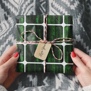 Peut inclure: Un cadeau emball&eacute; avec un motif de code binaire noir et vert, attach&eacute; avec de la ficelle rouge et blanche. Une petite &eacute;tiquette en bois avec "Love You!" est attach&eacute;e. Le cadeau est tenu par une personne.