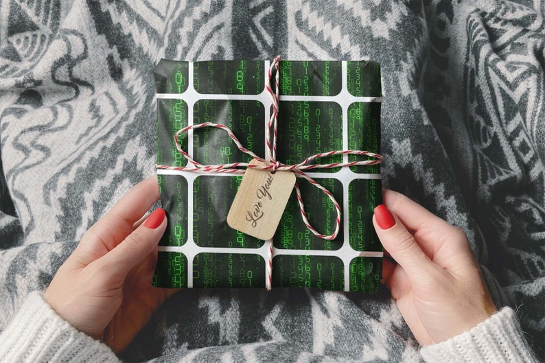 Peut inclure: Un cadeau emball&eacute; avec un motif de code binaire noir et vert, attach&eacute; avec de la ficelle rouge et blanche. Une petite &eacute;tiquette en bois avec "Love You!" est attach&eacute;e. Le cadeau est tenu par une personne.