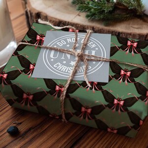 May include: A wrapped gift with green paper featuring a repeating pattern of black moths with pink bows. The gift is tied with twine and has a gray tag that reads "DO NOT OPEN UNTIL CHRISTMAS". The gift is on a wooden surface.