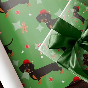 K&ouml;nnte beinhalten: Gr&uuml;nes Geschenkpapier und ein verpacktes Geschenk mit einer dunkelgr&uuml;nen Satinschleife. Das Papier zeigt ein sich wiederholendes Muster aus Dackeln mit Weihnachtsm&uuml;tzen, Sternen, Lebkuchenm&auml;nnern und Zuckerstangen. Das Geschenk ist bereit f&uuml;r die Feiertage.