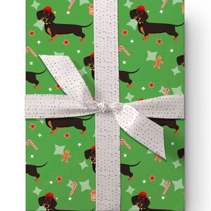 K&ouml;nnte beinhalten: Verpacktes Geschenk mit gr&uuml;nem Geschenkpapier mit einem sich wiederholenden Muster aus Dackeln mit Weihnachtsm&uuml;tzen, Zuckerstangen, Lebkuchenm&auml;nnern und Sternen. Ein wei&szlig;es Band mit grauen Punkten ist um das Geschenk gebunden.