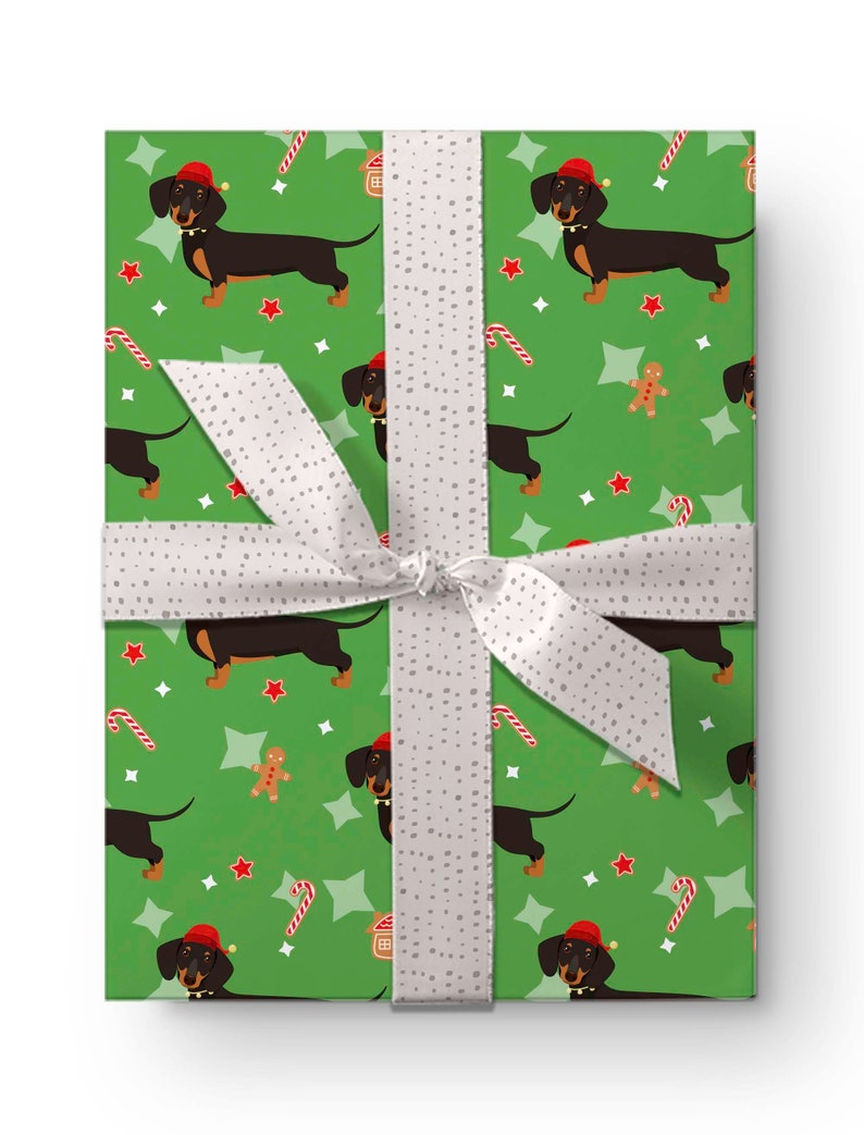 K&ouml;nnte beinhalten: Verpacktes Geschenk mit gr&uuml;nem Geschenkpapier mit einem sich wiederholenden Muster aus Dackeln mit Weihnachtsm&uuml;tzen, Zuckerstangen, Lebkuchenm&auml;nnern und Sternen. Ein wei&szlig;es Band mit grauen Punkten ist um das Geschenk gebunden.