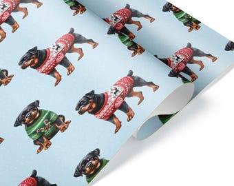 Può includere: Un rotolo di carta da regalo azzurro con un motivo ripetuto di cani Rottweiler. Alcuni cani indossano maglioni rossi con motivi a fiocchi di neve bianchi, mentre altri indossano maglioni verdi con disegni di bastoncini di zucchero.