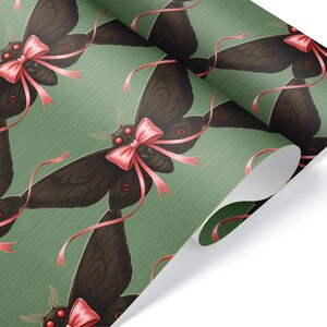 May include: A roll of wrapping paper featuring a repeating pattern of dark brown moths adorned with pink bows and ribbons. The moths have red eyes and are set against a green background with diagonal stripes.