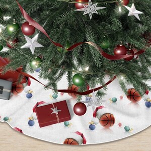 May include: A Christmas tree skirt with a basketball theme. The white skirt features basketballs, Santa hats, and ornaments. A decorated Christmas tree with red ribbon, ornaments, and star-shaped decorations sits on top.
