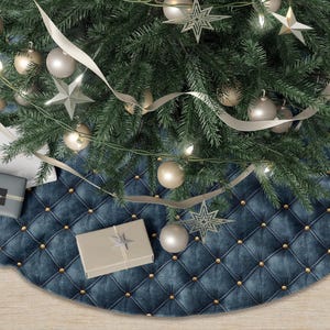 May include: A Christmas tree skirt with a tufted, navy blue velvet design and gold-colored button accents. The skirt is beneath a decorated Christmas tree with ornaments, lights, and ribbon. Several wrapped gifts are placed on the skirt.