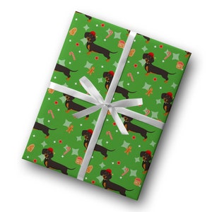 K&ouml;nnte beinhalten: Verpacktes Geschenk mit gr&uuml;nem Geschenkpapier mit einem sich wiederholenden Muster aus schwarzen Dackeln mit roten H&uuml;ten, Lebkuchenm&auml;nnern, Zuckerstangen und Sternen. Das Geschenk ist mit einem wei&szlig;en Band gebunden.