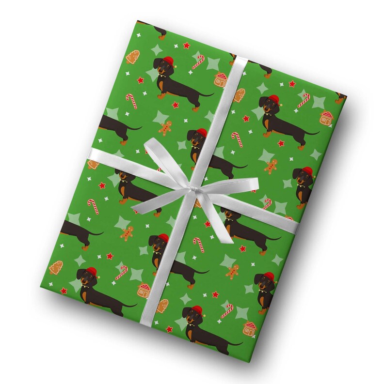 K&ouml;nnte beinhalten: Verpacktes Geschenk mit gr&uuml;nem Geschenkpapier mit einem sich wiederholenden Muster aus schwarzen Dackeln mit roten H&uuml;ten, Lebkuchenm&auml;nnern, Zuckerstangen und Sternen. Das Geschenk ist mit einem wei&szlig;en Band gebunden.