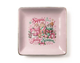 Può includere: Un piattino quadrato in ceramica rosa chiaro con bordo dorato. Il centro presenta una colorata illustrazione natalizia con tre bevande, un pupazzo di neve e la scritta Sippin' into a Jolly Season in caratteri rossi e verdi.