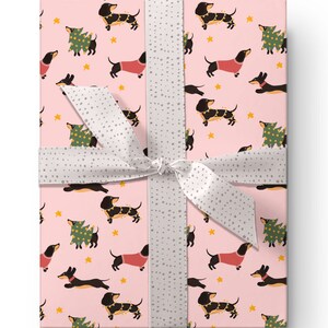Op de afbeelding: Roze cadeaupapier met een herhalend patroon van zwarte teckels die rode truien en kerstverlichting dragen, en groene kerstboom met verlichting. Het papier is vastgebonden met een wit lint met grijze stippen.