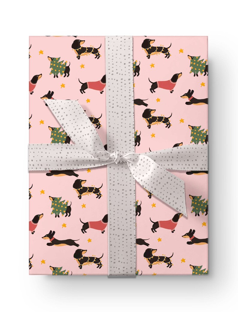 Op de afbeelding: Roze cadeaupapier met een herhalend patroon van zwarte teckels die rode truien en kerstverlichting dragen, en groene kerstboom met verlichting. Het papier is vastgebonden met een wit lint met grijze stippen.
