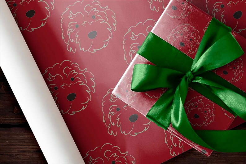 May include: A red gift box with a green satin ribbon bow sits atop red wrapping paper patterned with light green dog faces. A roll of white wrapping paper is visible on the left.