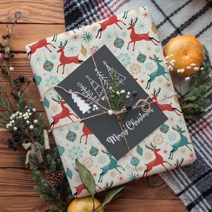 May include: A wrapped gift with festive reindeer and ornament pattern paper. A black card with "Merry Christmas" is tied with twine. The gift is decorated with greenery and a tangerine on a wooden surface.