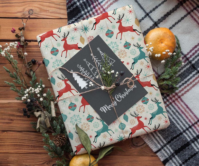 May include: A wrapped gift with festive reindeer and ornament pattern paper. A black card with "Merry Christmas" is tied with twine. The gift is decorated with greenery and a tangerine on a wooden surface.
