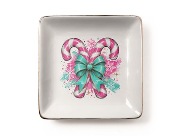 Può includere: Un piattino quadrato in ceramica bianca con un bordo dorato metallizzato. Il centro presenta un'illustrazione grafica di due bastoncini di zucchero incrociati a strisce rosa e bianche, legati da un grande fiocco turchese e fiocchi.