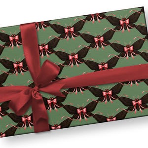 May include: A wrapped gift box featuring a repeating pattern of black moths with pink bows on a green background. The box is tied with a wide, dark red satin ribbon and bow, creating a festive presentation.