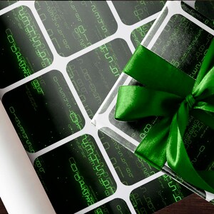 Peut inclure: Un cadeau emball&eacute; avec un ruban vert en satin. Le papier d'emballage a un fond noir avec un motif de grille blanche et des motifs verts ressemblant &agrave; des codes num&eacute;riques. Un rouleau de papier d'emballage assorti est visible.