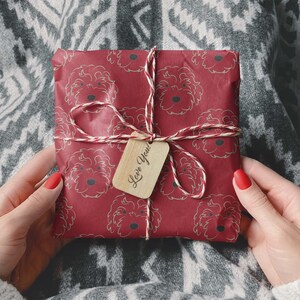 May include: A wrapped gift with red paper featuring a repeating pattern of dog faces. The gift is tied with twine and a small wooden tag that says "Love You!". The gift is held by a person wearing a white sweater.