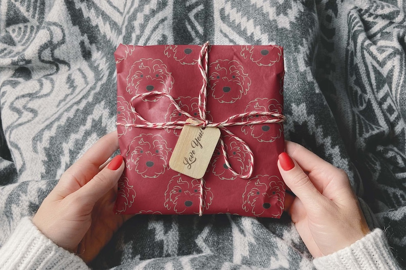 May include: A wrapped gift with red paper featuring a repeating pattern of dog faces. The gift is tied with twine and a small wooden tag that says "Love You!". The gift is held by a person wearing a white sweater.