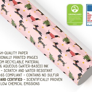 Op de afbeelding: Roze inpakpapier met een herhalend patroon van zwarte en bruine teckels die rode truien dragen en kerstboompjes vasthouden. Het papier is gemaakt van recyclebaar materiaal en is GreenGuard-gecertificeerd.