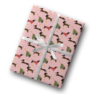 Op de afbeelding: Roze cadeaupapier met een herhalend patroon van zwarte en bruine teckels die rode truien en groene kerstboom dragen. Een wit lint is om het papier gebonden.