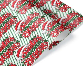 Può includere: Un rotolo di carta da regalo con un motivo ripetuto di testo rosso e verde che recita Santa's Favorite Stylist, accentuato da cappelli di Babbo Natale rossi e foglie di agrifoglio verdi su uno sfondo a sottili strisce diagonali verdi.