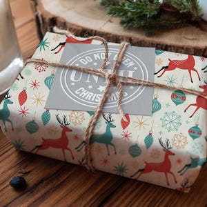 May include: A wrapped gift with festive Christmas-themed paper featuring red and teal reindeer, ornaments, and snowflakes. The package is tied with twine and a gray tag that reads "DO NOT OPEN UNTIL CHRISTMAS".