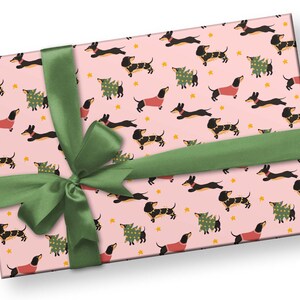Op de afbeelding: Roze cadeaupapier met een herhalend patroon van zwarte en bruine teckels die rode truien en kerstboomkostuums dragen. Een groen lint is in een strik om het papier gebonden.