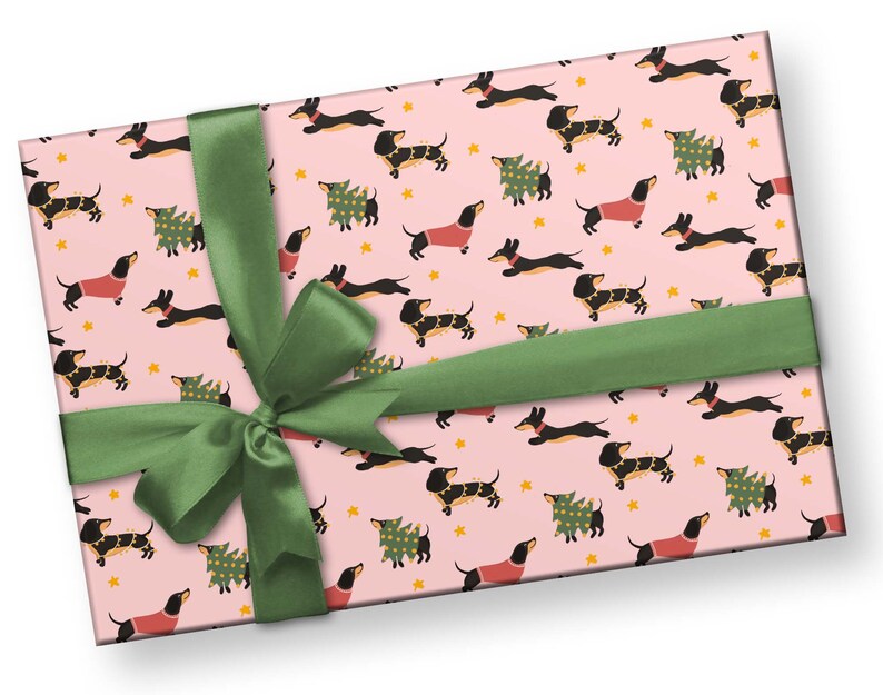 Op de afbeelding: Roze cadeaupapier met een herhalend patroon van zwarte en bruine teckels die rode truien en kerstboomkostuums dragen. Een groen lint is in een strik om het papier gebonden.
