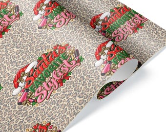 Può includere: Carta da regalo con un motivo ripetuto su sfondo leopardato e il testo Santa's Favorite Stylist. Il design include un cappello da Babbo Natale rosso, un asciugacapelli rosa, forbici dorate e una spazzola per capelli rotonda con dettagli leopardati.