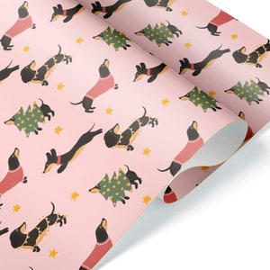 Op de afbeelding: Roze cadeaupapier met een herhalend patroon van zwarte teckels die rode truien en kerstverlichting dragen, en groene kerstboom met gele lampjes.