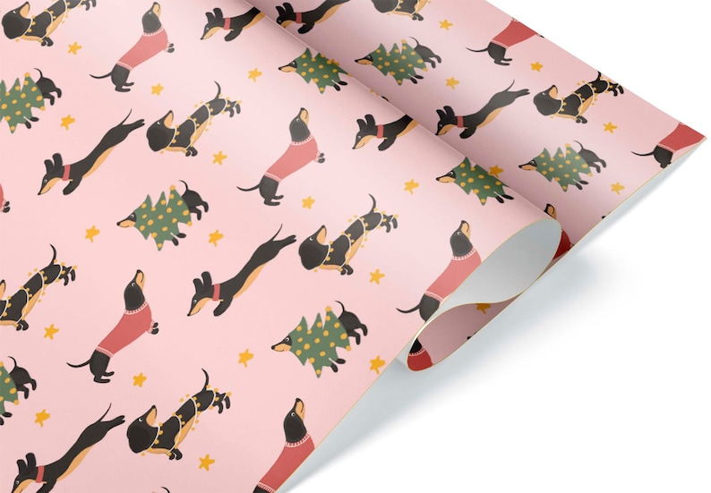 Op de afbeelding: Roze cadeaupapier met een herhalend patroon van zwarte teckels die rode truien en kerstverlichting dragen, en groene kerstboom met gele lampjes.