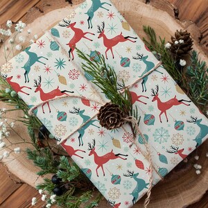 May include: Two wrapped gifts with festive wrapping paper featuring red and teal reindeer, snowflakes, and ornaments. The gifts are tied with twine and decorated with pine, berries, and a pinecone, set on a wooden surface.