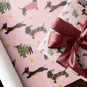 Op de afbeelding: Roze cadeaupapier met een herhalend patroon van zwarte teckels die kerstboom en truien dragen. Een bordeauxrood lint is gebonden om een ​​cadeaudoos die in hetzelfde papier is gewikkeld.