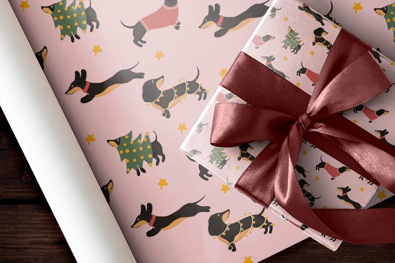 Op de afbeelding: Roze cadeaupapier met een herhalend patroon van zwarte teckels die kerstboom en truien dragen. Een bordeauxrood lint is gebonden om een ​​cadeaudoos die in hetzelfde papier is gewikkeld.