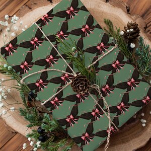 May include: A wrapped gift with green paper featuring a repeating pattern of black butterflies with pink bows. The gift is tied with twine and decorated with pine, berries, and a pinecone, set on a wooden surface.