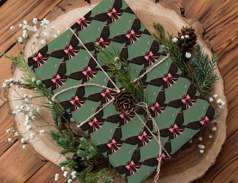 May include: A wrapped gift with green paper featuring a repeating pattern of black butterflies with pink bows. The gift is tied with twine and decorated with pine, berries, and a pinecone, set on a wooden surface.