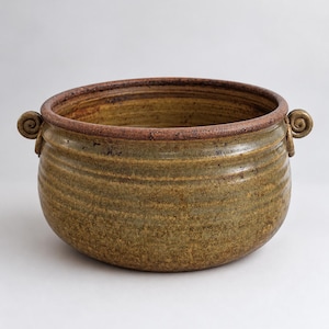 May include: A round, hand-thrown ceramic bowl featuring a mottled olive-green and brown glaze with horizontal ridges along the exterior. The bowl has a thick, dark brown rim and two small, decorative spiral-shaped handles on opposite sides.