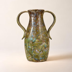 May include: A ceramic vase featuring two curved handles and a flared rim. The surface displays a mottled glaze with blended tones of earthy brown, moss green, and streaks of light blue, creating a textured appearance across the entire body.
