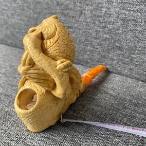 Può includere: Una statuetta scolpita color beige di una creatura mitica con corpo di pesce e braccia umane, che si tiene il viso. La statuetta ha una coda arancione e un'apertura circolare. Un metro a nastro è accanto.