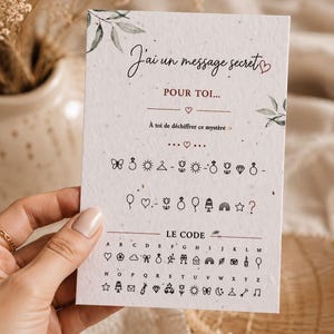 Peut inclure: Une carte rectangulaire blanche avec le texte J'ai un message secret POUR TOI, suivi d'une série de petites icônes noires représentant un code secret, avec une clé de décodage alphabétique complète en bas associant lettres et symboles.
