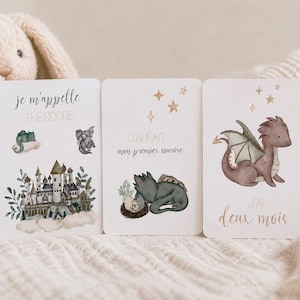 Peut inclure: Trois cartes étapes rectangulaires illustrées d'aquarelles de dragons et de texte. Les cartes affichent les phrases je m'appelle THEODORE, J'AI FAIT mon premier sourire et J'AI deux mois, accompagnées de dessins d'un château et de dragons.