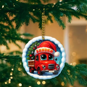 May include: A round Christmas ornament featuring a red truck with big eyes and a Santa hat that says "Sandra". The ornament has a snowy border and a Christmas tree in the background. The ornament is hanging from a gold string.